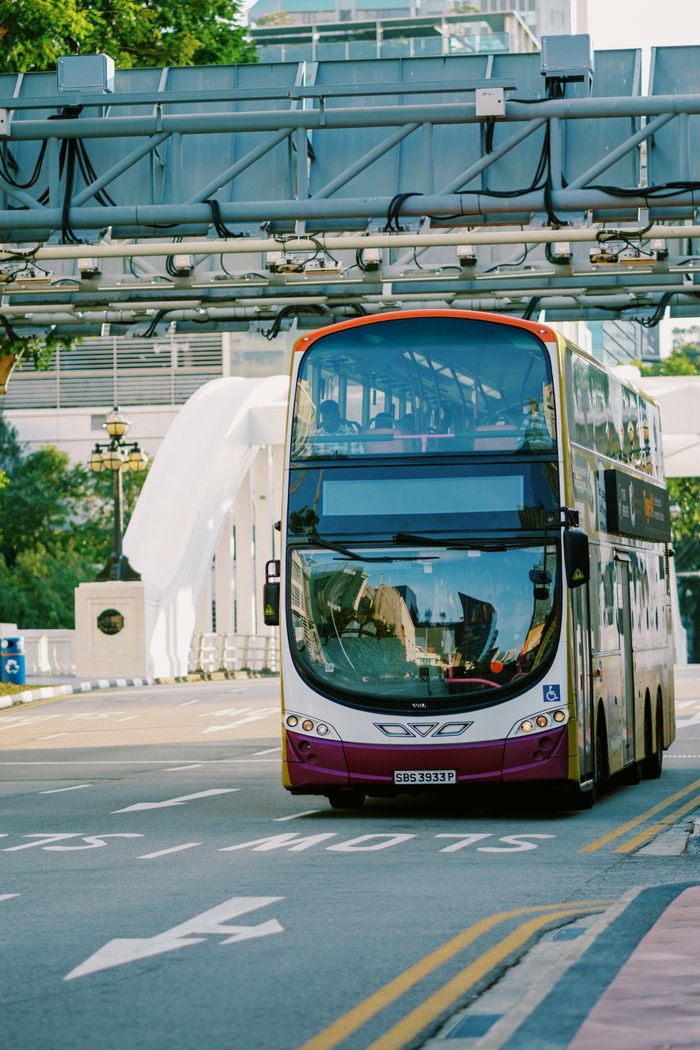 A double-decker bus drives under a sky bridge on a city street during the day.