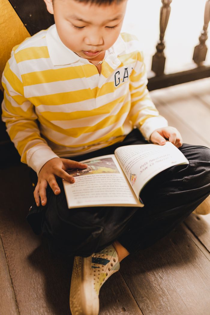 Young Asian boy in a yellow striped shirt focused on reading a book indoors.